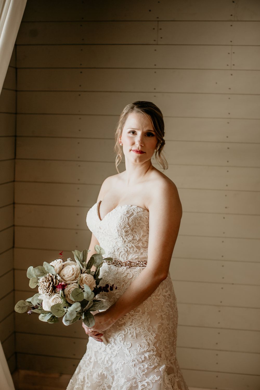 Bride in strapless lace mermaid gown with crystal belt holding eucalyptus and rose bouquet.