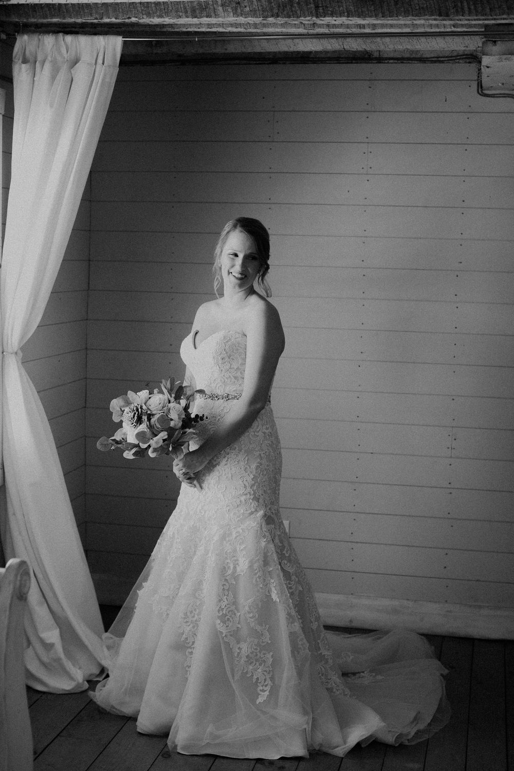 Black and white portrait of smiling bride in lace mermaid gown holding bouquet by curtain.