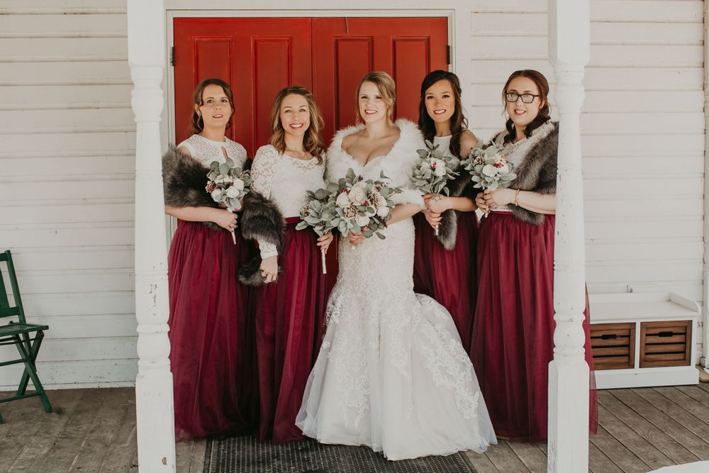 Bride with four bridesmaids in burgundy gowns holding bouquets in front of red barn door.