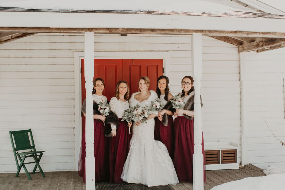 Bride and four bridesmaids in burgundy dresses posing outside white barn with red door.