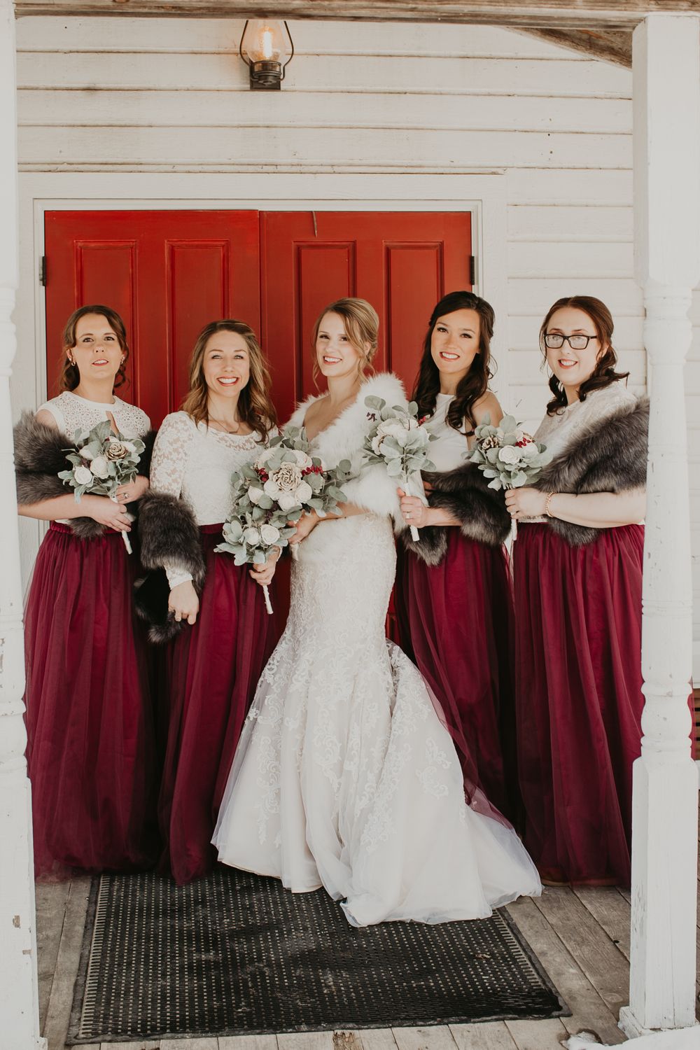 Bride in white lace gown with fur wrap poses with four bridesmaids in burgundy dresses before red barn doors.