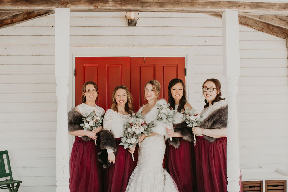 Bride flanked by four bridesmaids in burgundy gowns with fur wraps holding floral bouquets.
