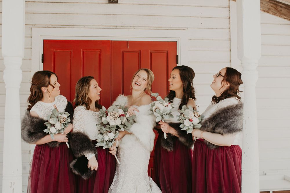 Laughing bride and bridesmaids in burgundy dresses with fur stoles hold bouquets before a red door.