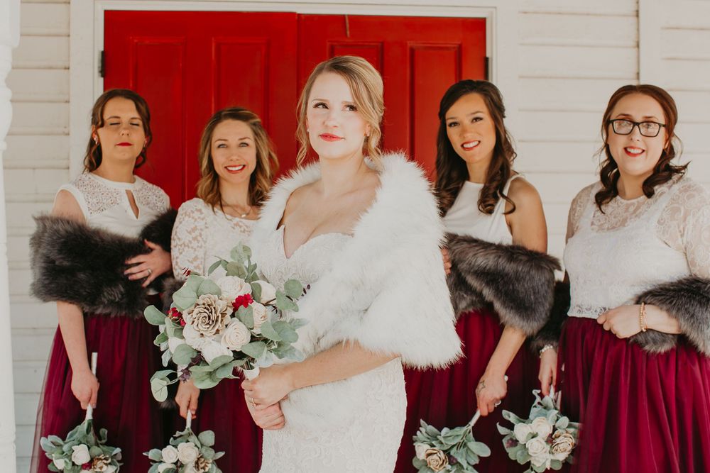 Blonde bride in white fur wrap stands with four bridesmaids in burgundy gowns holding winter bouquets.