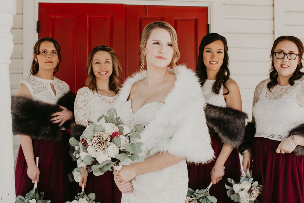 Bride in white fur stole poses with smiling bridesmaids in burgundy dresses holding floral bouquets.