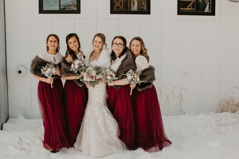 Bridal party in burgundy dresses and fur wraps poses together in the snow holding winter bouquets.