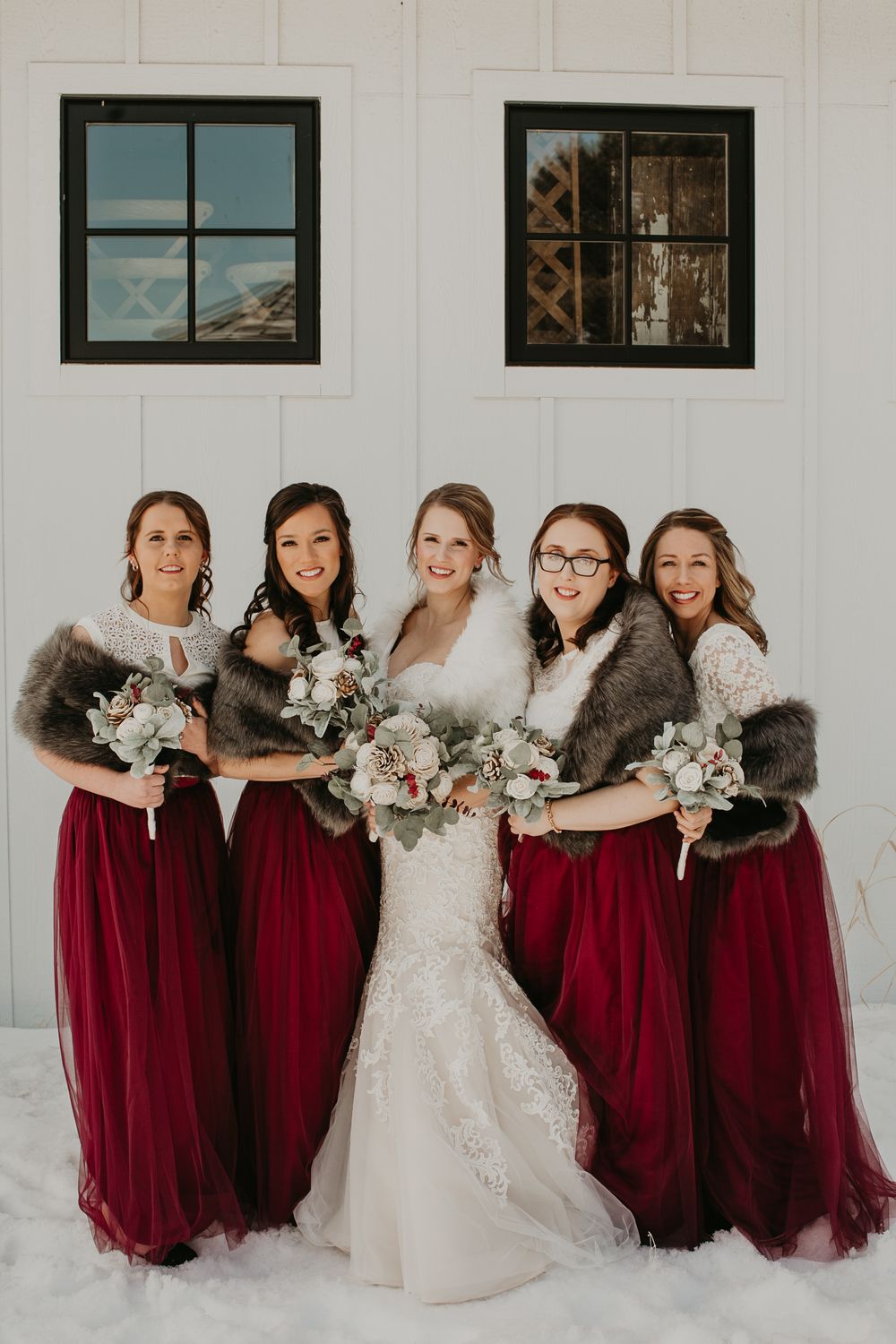 Winter wedding party with bride in lace gown and bridesmaids in burgundy dresses pose in snow.