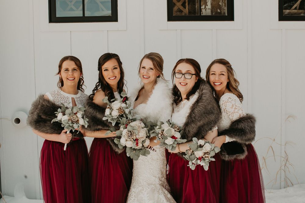 Smiling bride and bridesmaids in burgundy gowns with gray fur stoles hold bouquets in winter setting.