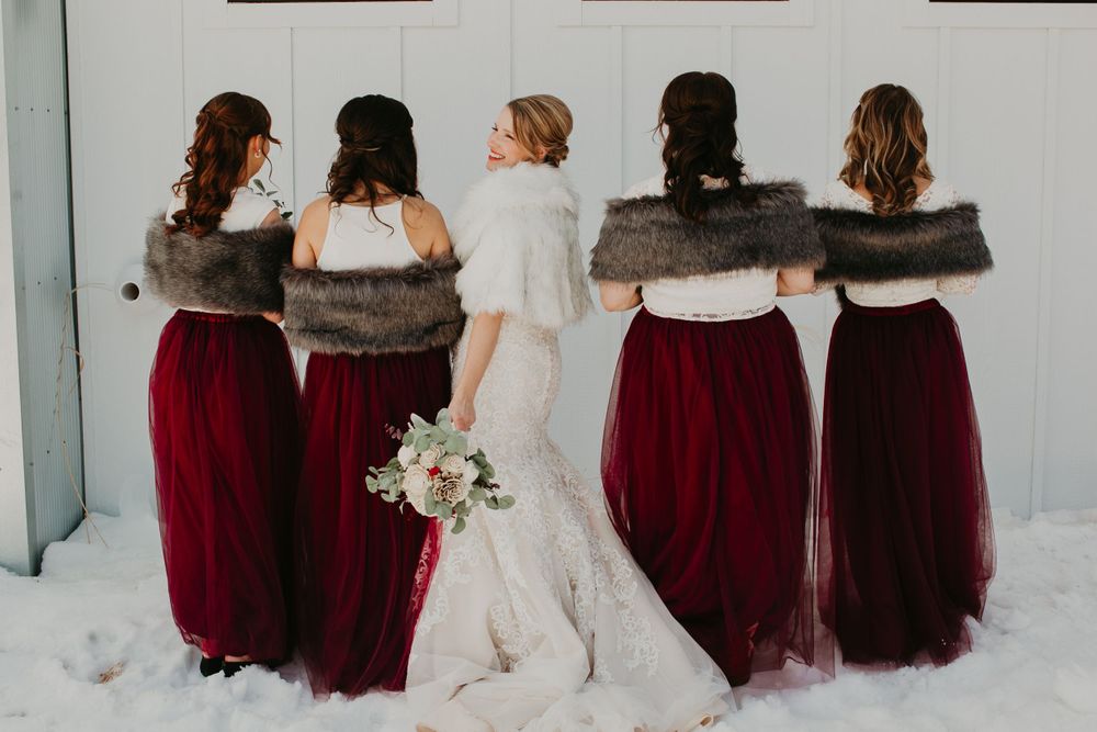 Bride and bridesmaids in burgundy dresses with fur wraps pose from behind in snowy winter scene.