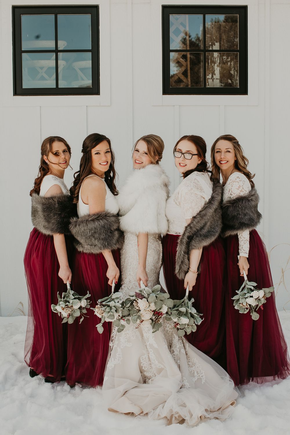 Bride in lace gown with fur wrap laughs with bridesmaids in burgundy dresses in winter snow.