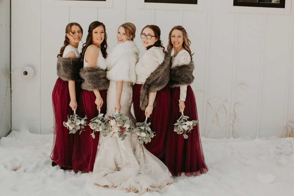 Bride and four bridesmaids in burgundy gowns with fur wraps pose in snow for a winter wedding.
