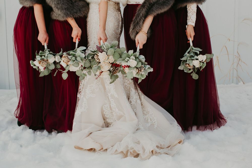 Close-up of bride and bridesmaids in burgundy dresses displaying elegant winter floral bouquets in snow.