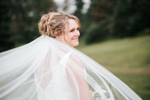 A bride smiles joyfully as her veil flows in the wind against a natural outdoor backdrop.