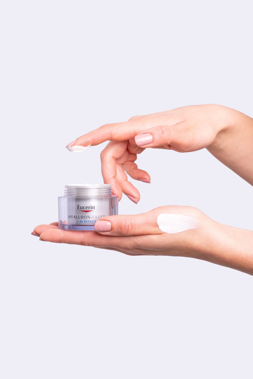 Hands holding and applying skincare cream from a small jar against white background.