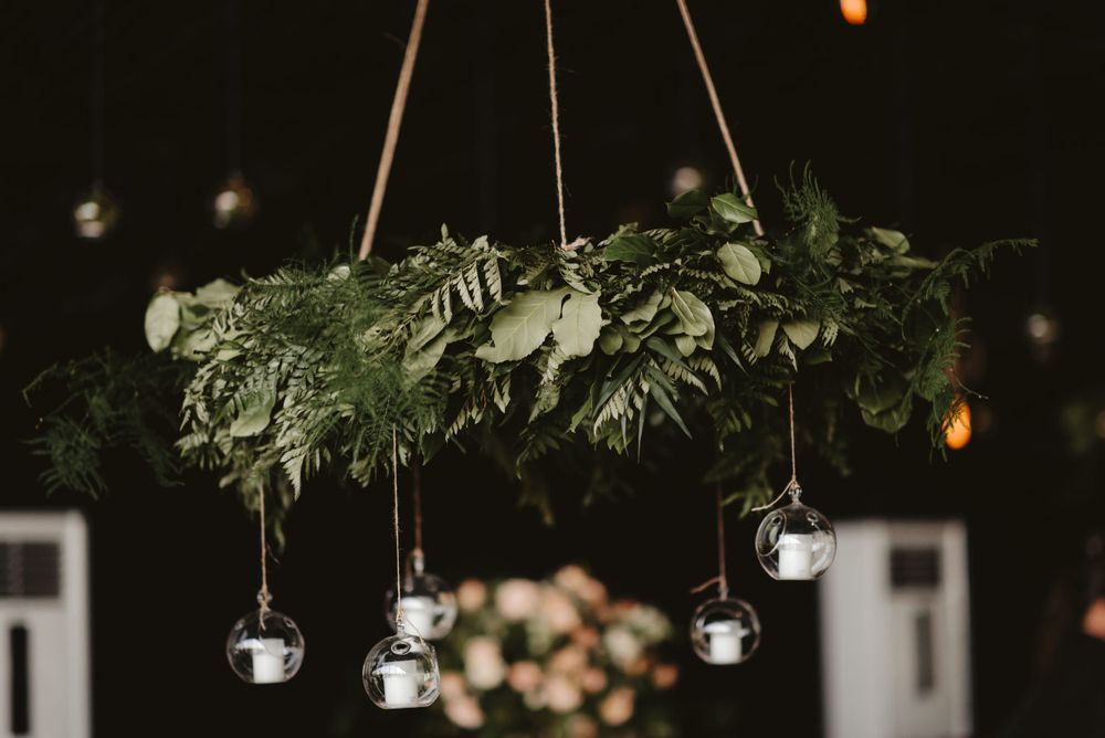 Rustic wedding decor featuring a suspended greenery chandelier with hanging glass tea light holders against a dark background.