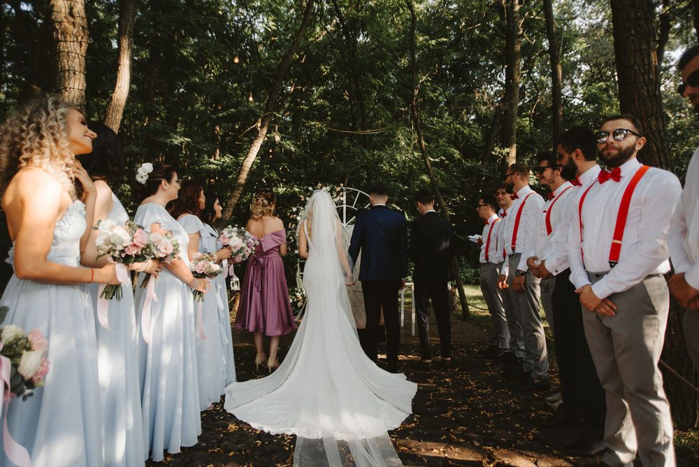 Outdoor forest wedding ceremony with wedding party in light blue dresses and red suspenders.