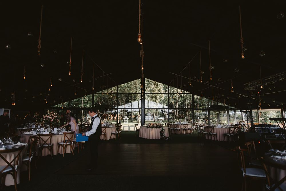 Dark reception venue with draped lighting and elegant table settings under a transparent tent ceiling.