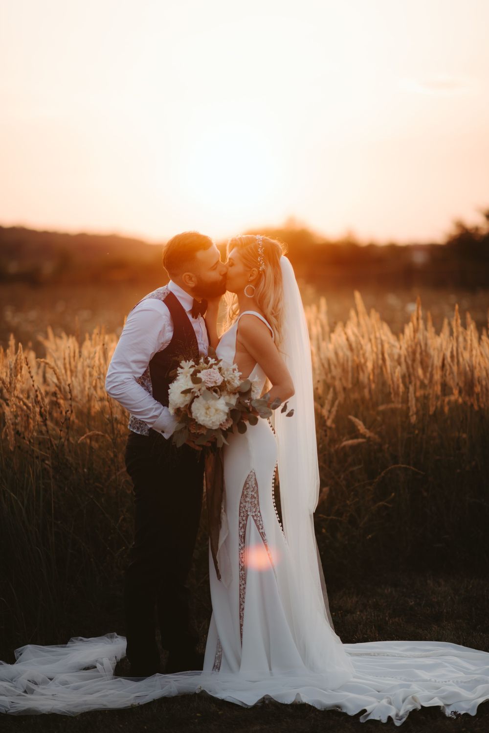 Romantic sunset wedding portrait in golden field with flowing white dress and bouquet.