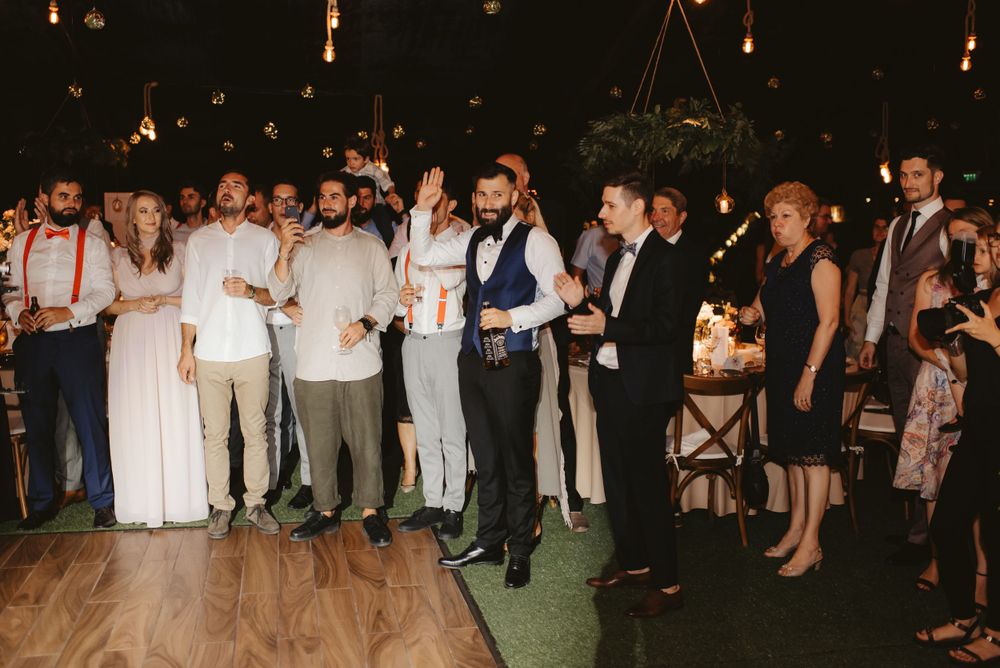 Wedding guests in formal attire gather on a dance floor under hanging lights.