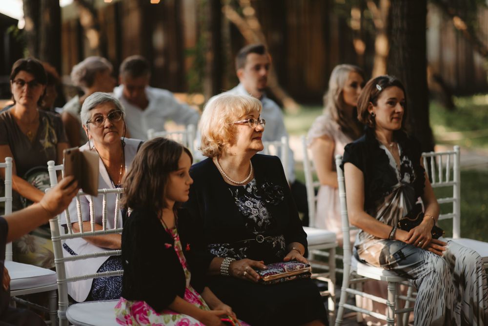 Guests seated in white chairs at outdoor wedding ceremony as sunlight filters through trees.
