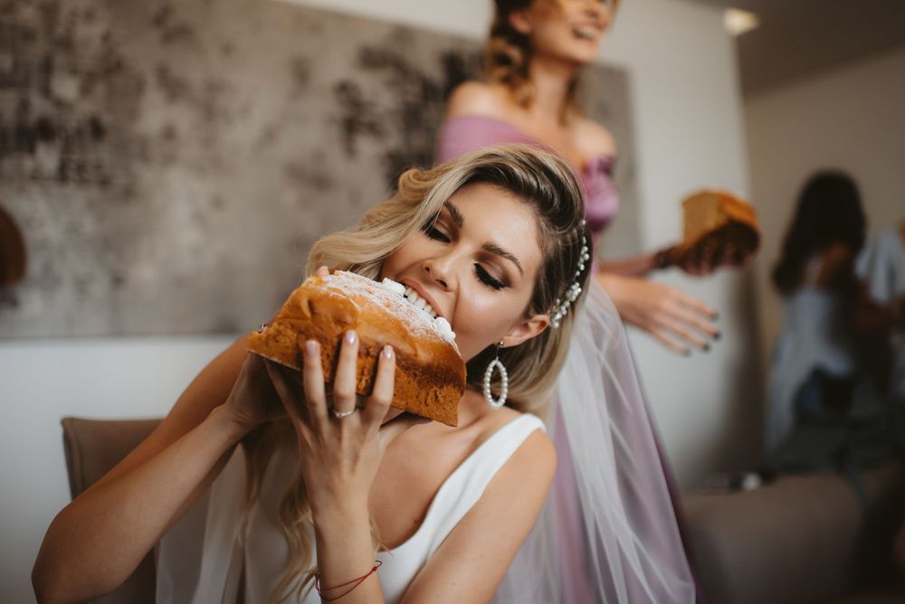 Candid moment of a bride taking a bite of food during wedding preparations with bridesmaids in background.