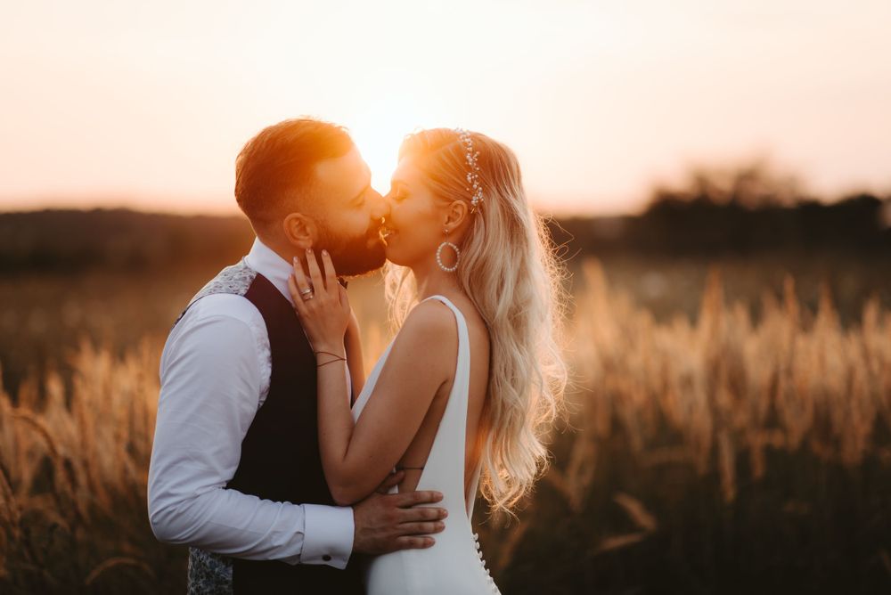 Newlyweds kiss in sunlit field with warm sunset glow.