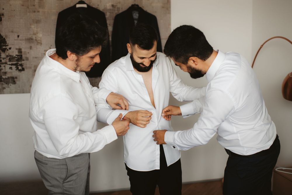 Groomsmen helping with wedding preparation as they assist with buttoning a white dress shirt.