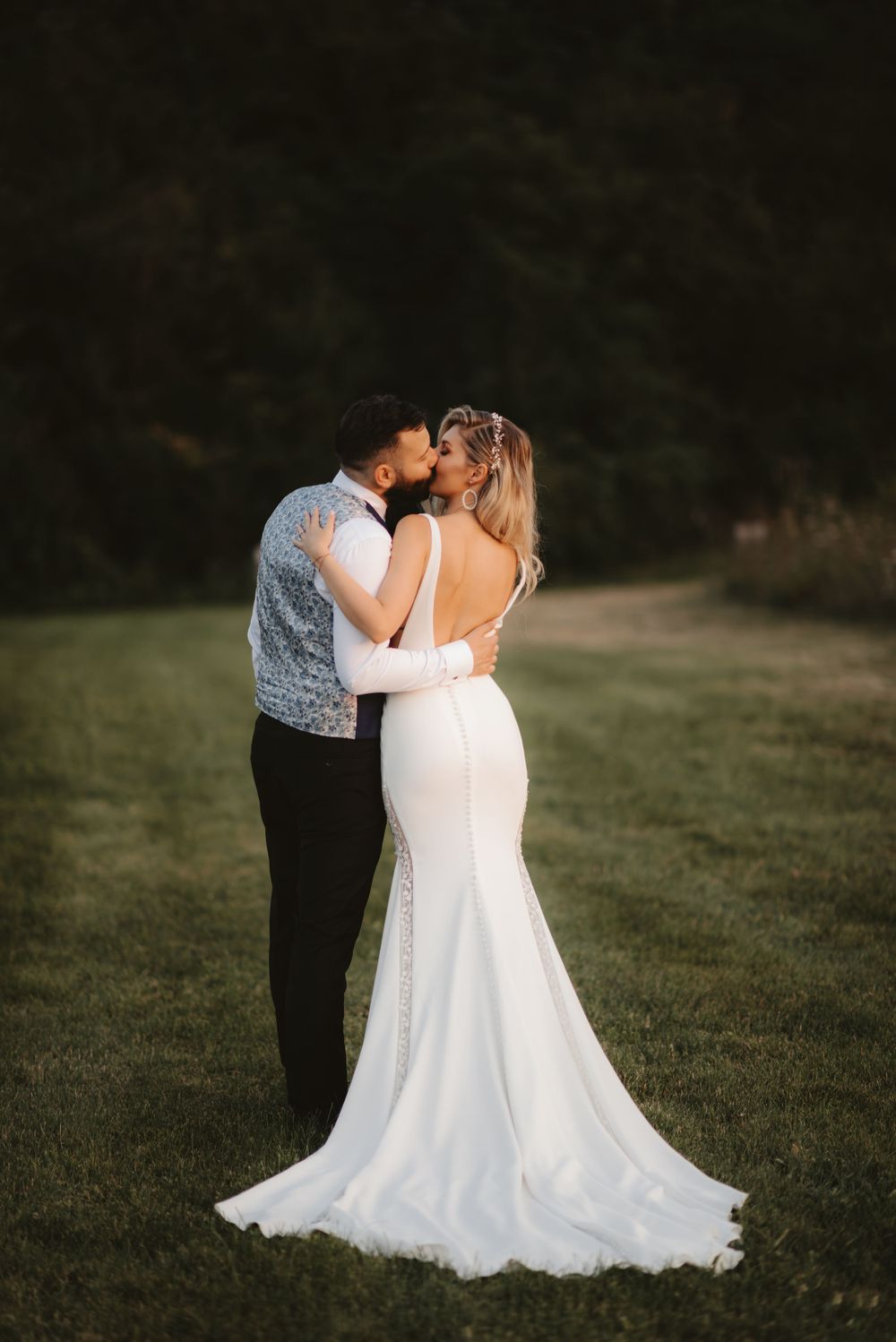 Wedding couple embraces on grassy hillside in elegant dress and vest.