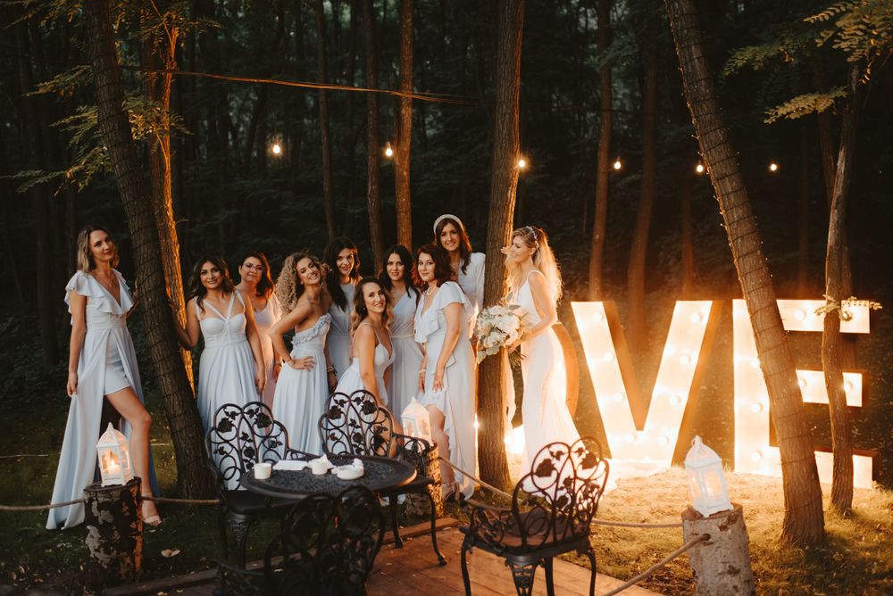 Bridesmaids in light dresses pose beside illuminated LOVE letters at dusk in a forest setting.