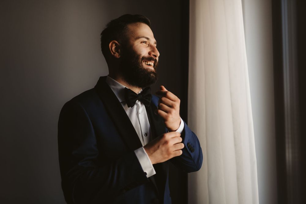 A well-dressed person in a suit adjusts their bowtie while standing by a window.