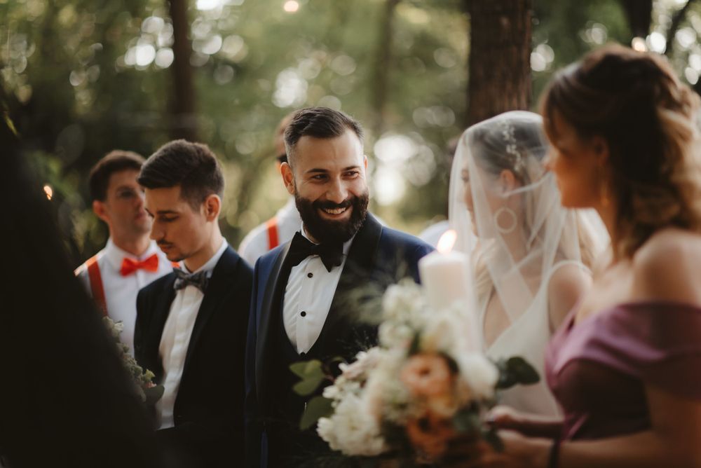Wedding party gathers outdoors in formal attire with bride in white veil among trees during golden hour.