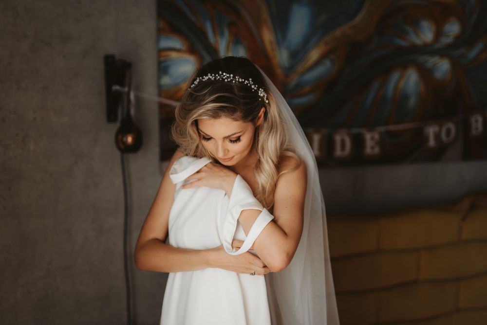 Bride in white dress and veil hugs herself while looking down in an intimate pre-wedding moment.