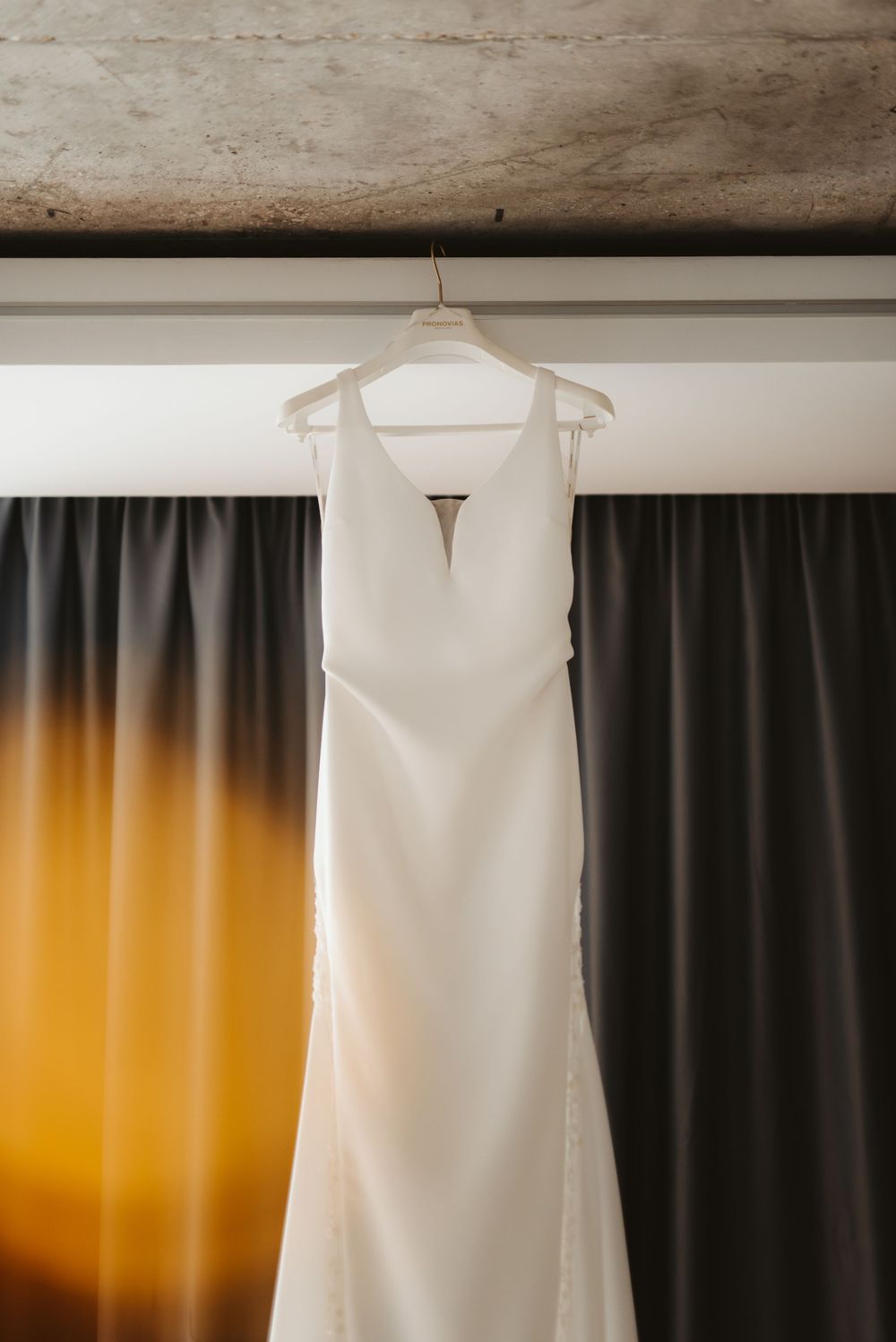 Elegant white wedding dress with plunging neckline hanging against dark curtains.