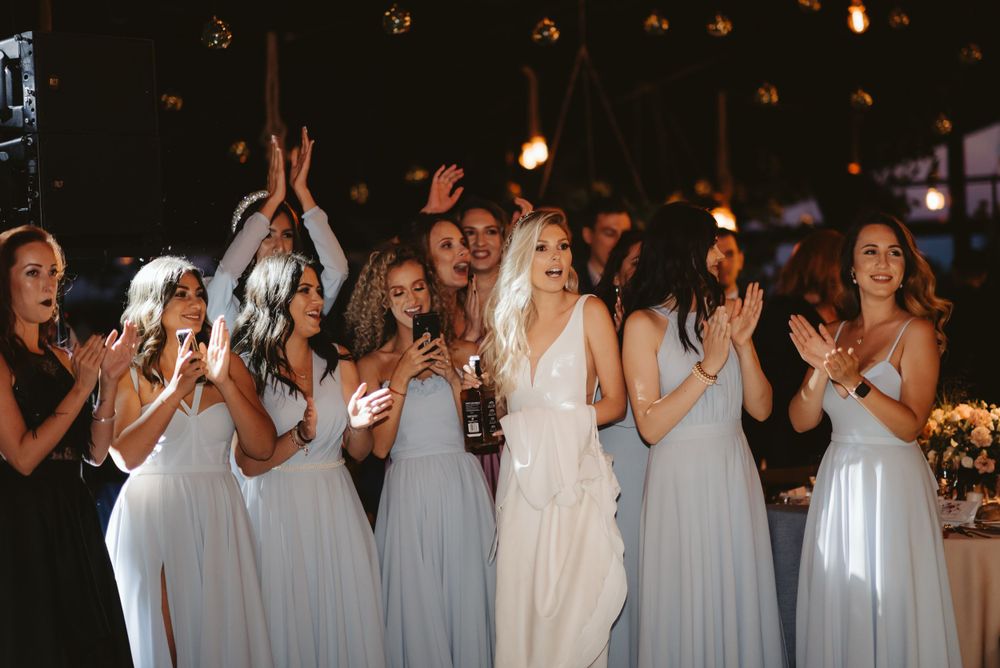 Wedding party in flowing white dresses celebrates during an indoor reception with ambient lighting.