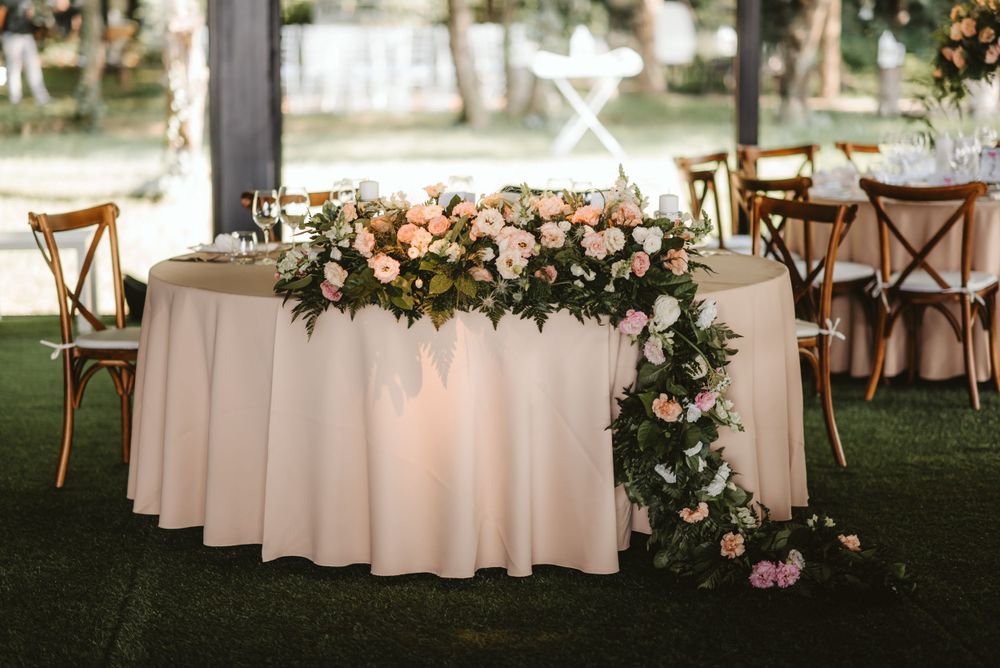 Romantic sweetheart table with cascading floral arrangement and blush tablecloth at outdoor wedding reception.