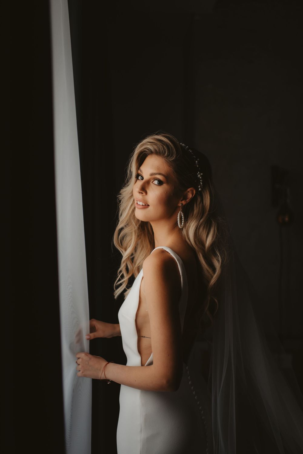 A bride in a sleek white wedding dress poses by a window, with soft moody lighting creating a dramatic silhouette.