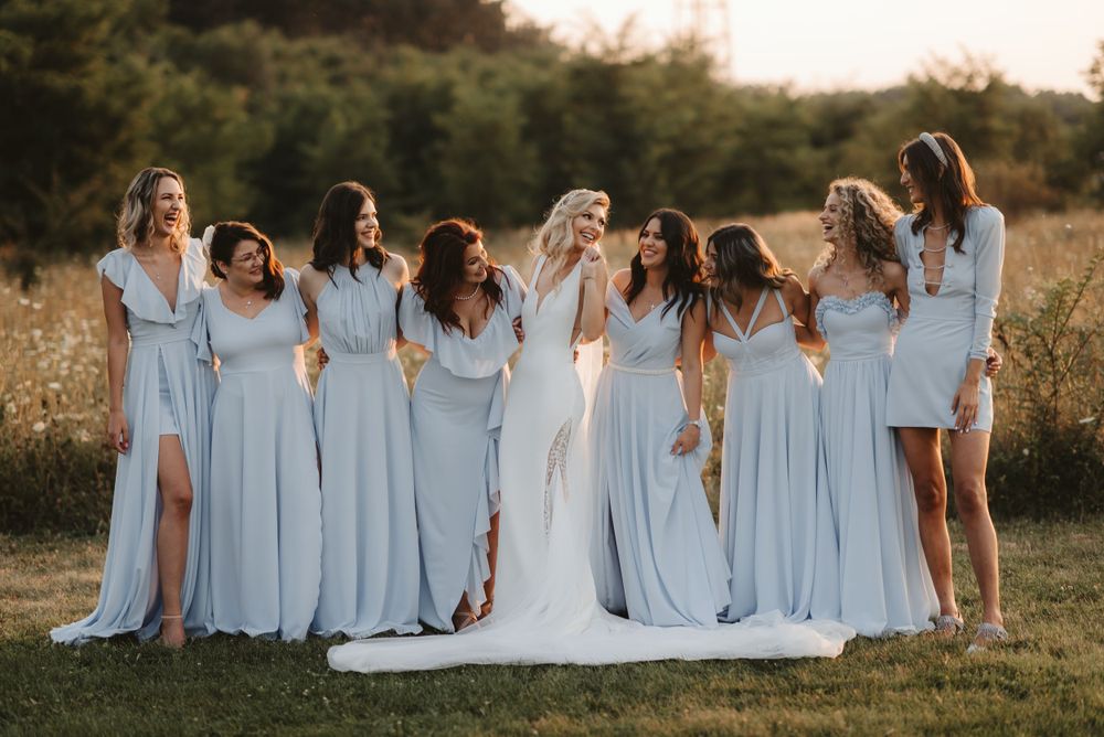 Bridesmaids in flowing light blue dresses pose together with bride in white gown during sunset outdoor photoshoot.