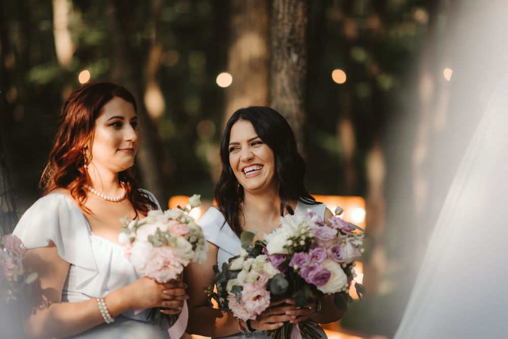 Two bridesmaids holding floral bouquets share a joyful moment in natural outdoor lighting.