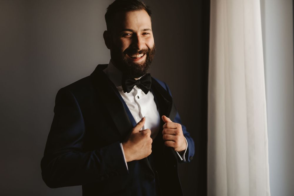 Man in formal tuxedo and bowtie adjusting his jacket by window light.
