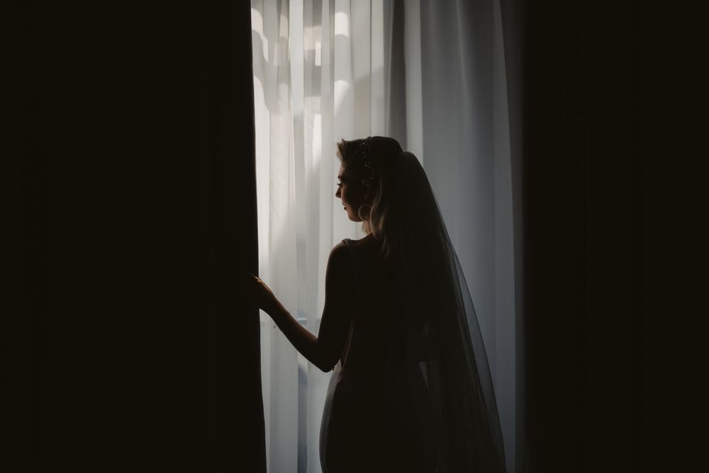 A silhouetted figure stands by a white curtained window creating a dramatic shadowy bridal portrait.