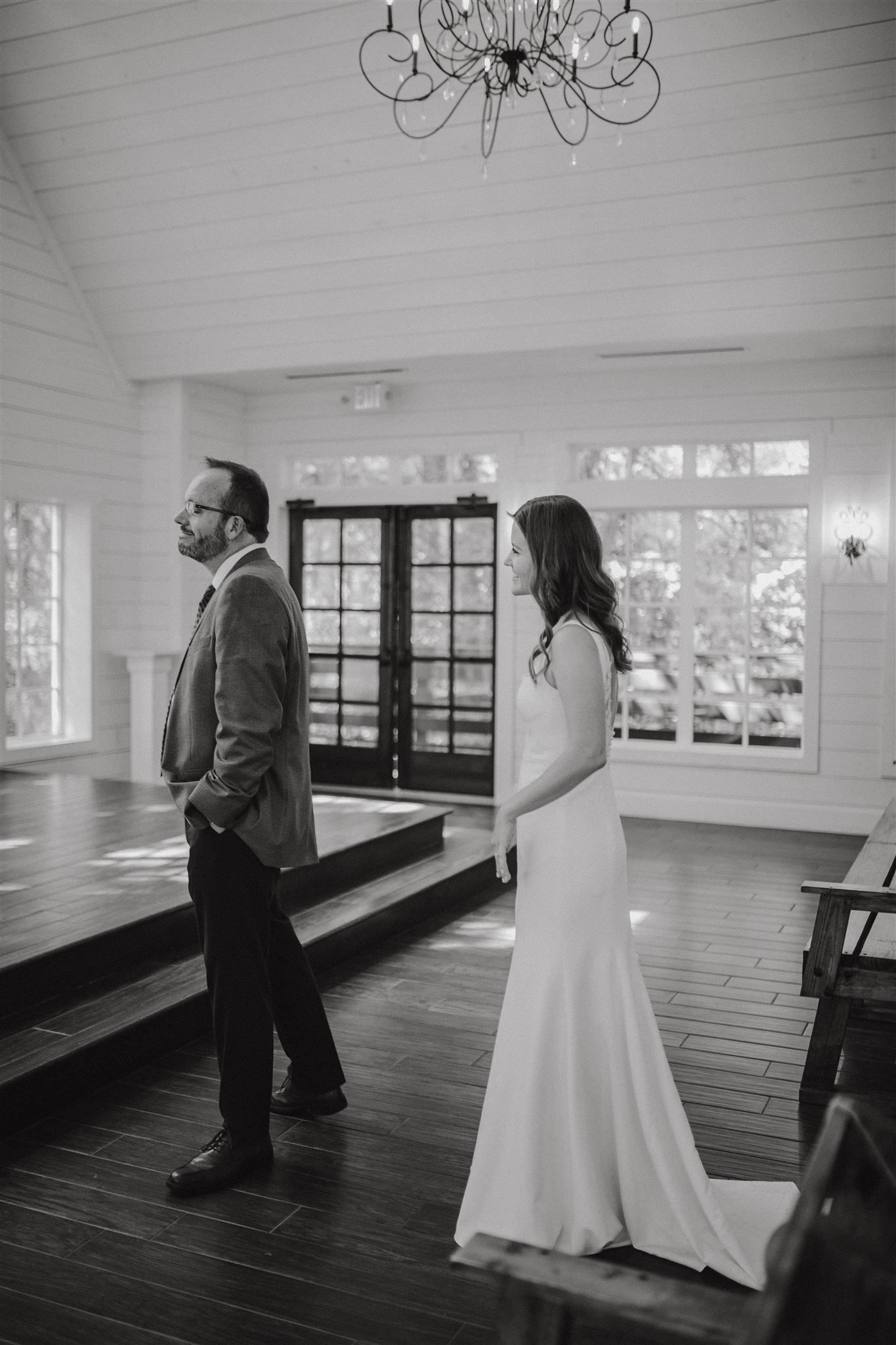 A couple in wedding attire standing apart in a bright white chapel with hardwood floors.