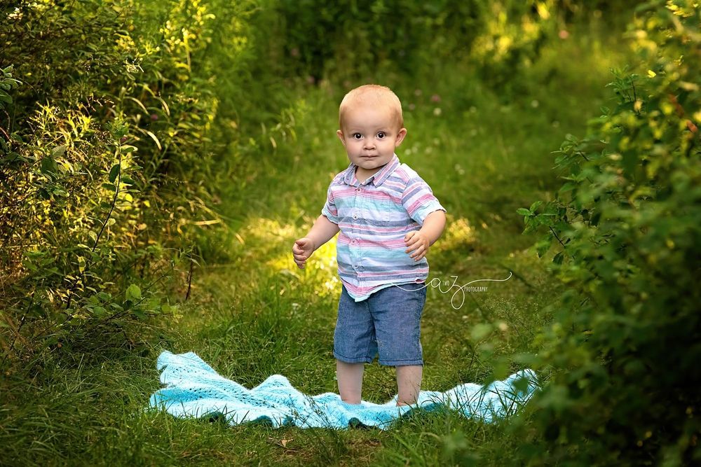 Toddler in striped shirt and shorts stands on blue fabric amid green foliage.