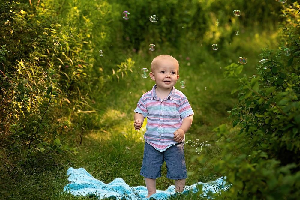 A small child in a striped shirt and blue shorts stands on a blue blanket in a lush green outdoor setting.
