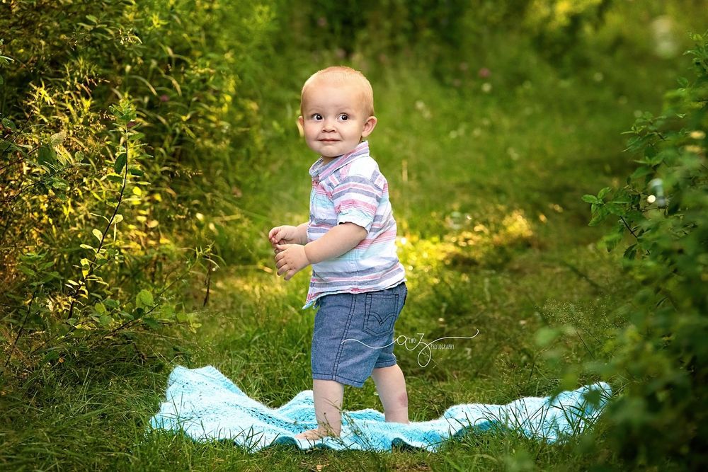 Smiling toddler in striped shirt poses on blue fabric in sunlit garden setting.