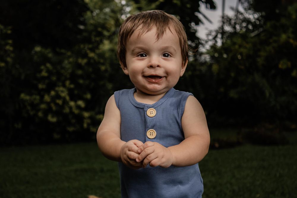 Toddler in blue sleeveless romper smiling in outdoor garden setting.
