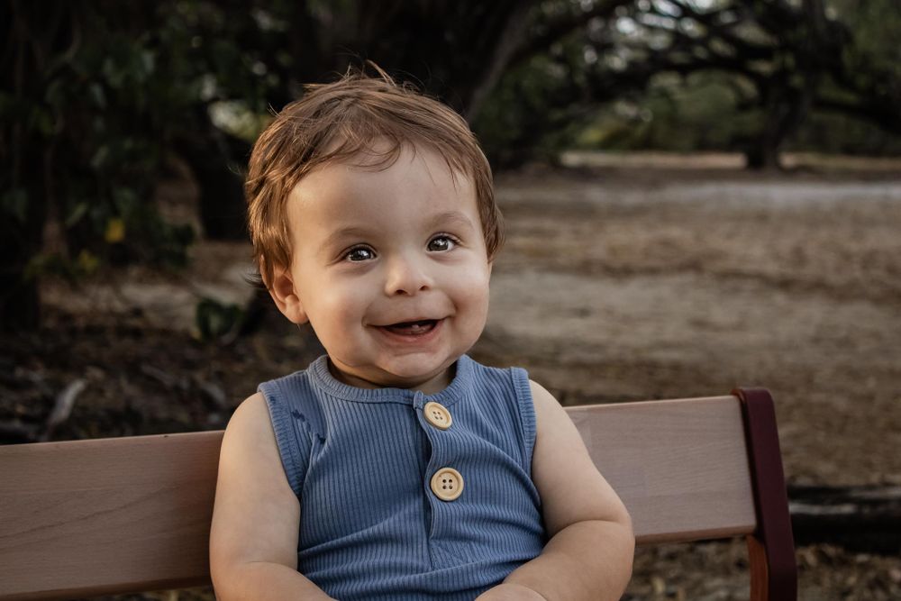 Happy toddler wearing blue button romper sitting on wooden bench outdoors.
