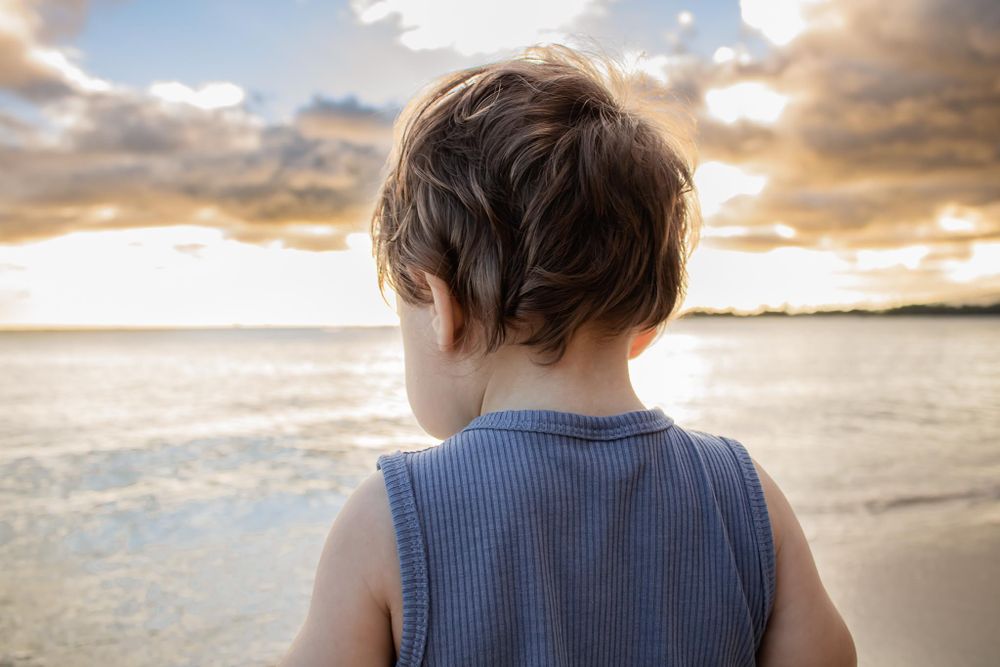 Back view of child in blue tank top watching sunset over ocean.