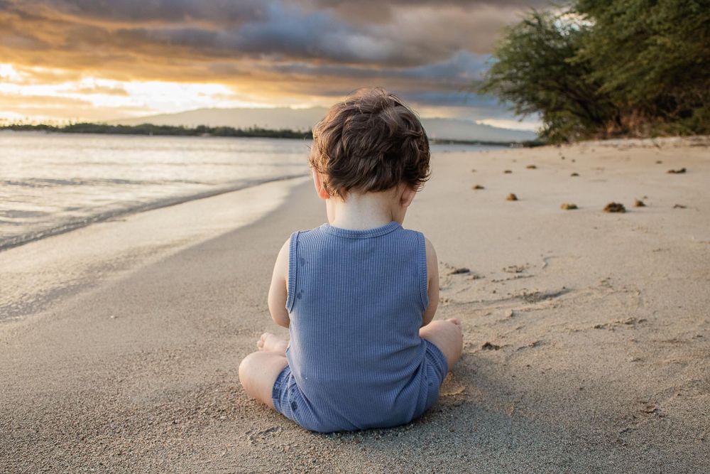 Child in blue romper sits alone on beach at sunset.