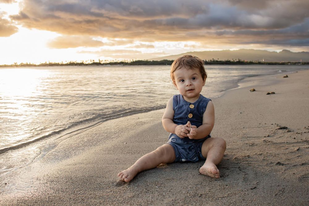 Toddler in blue outfit sits on sandy beach during golden sunset.