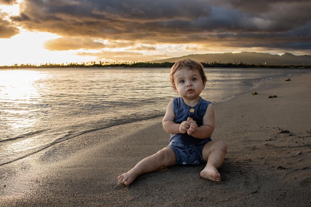 Child sits on beach sand during dramatic sunset with clouds.